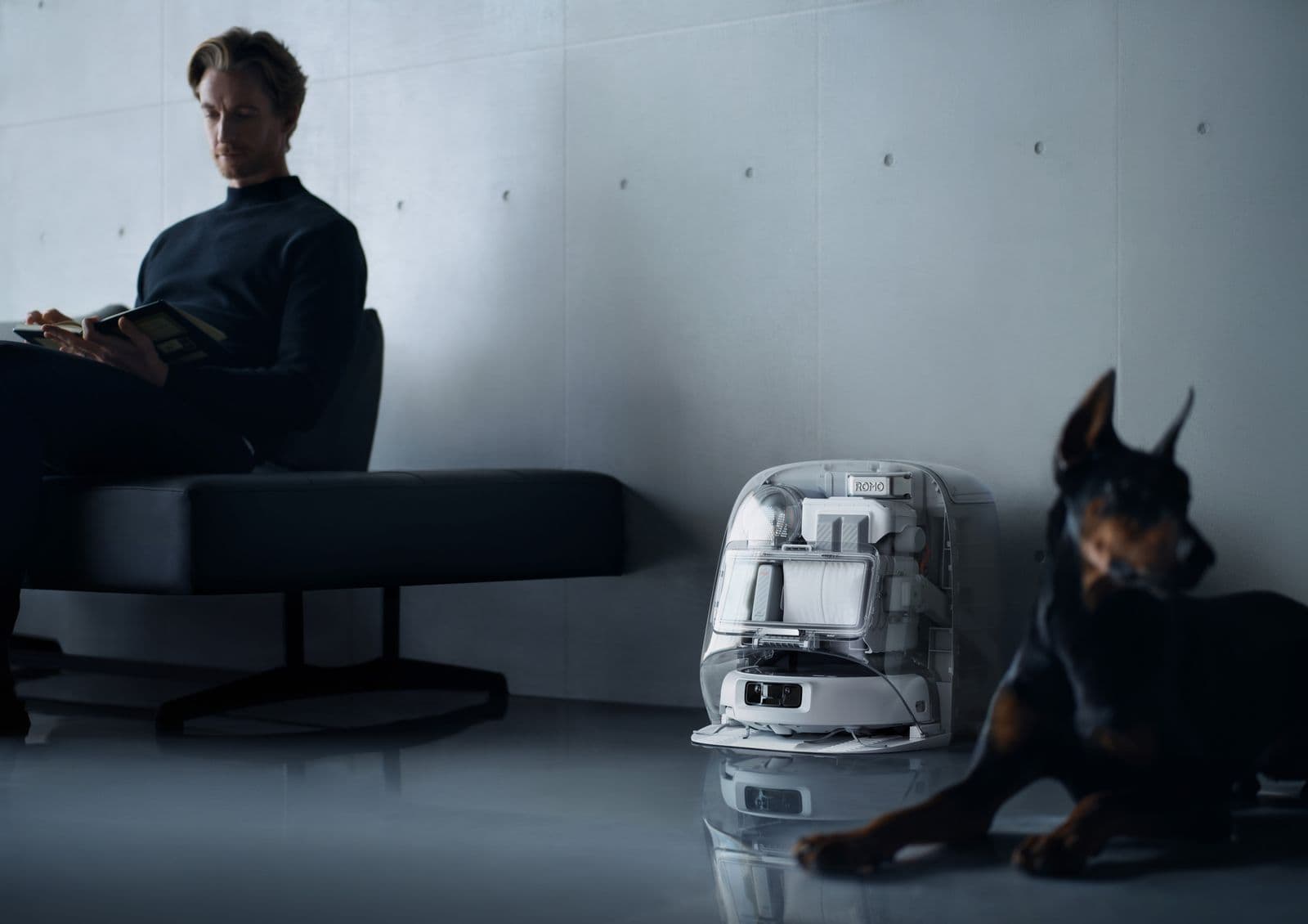 En man sitter och läser en bok på en modern soffa medan en hund ligger på golvet bredvid en kompakt robot i ett minimalistiskt rum.