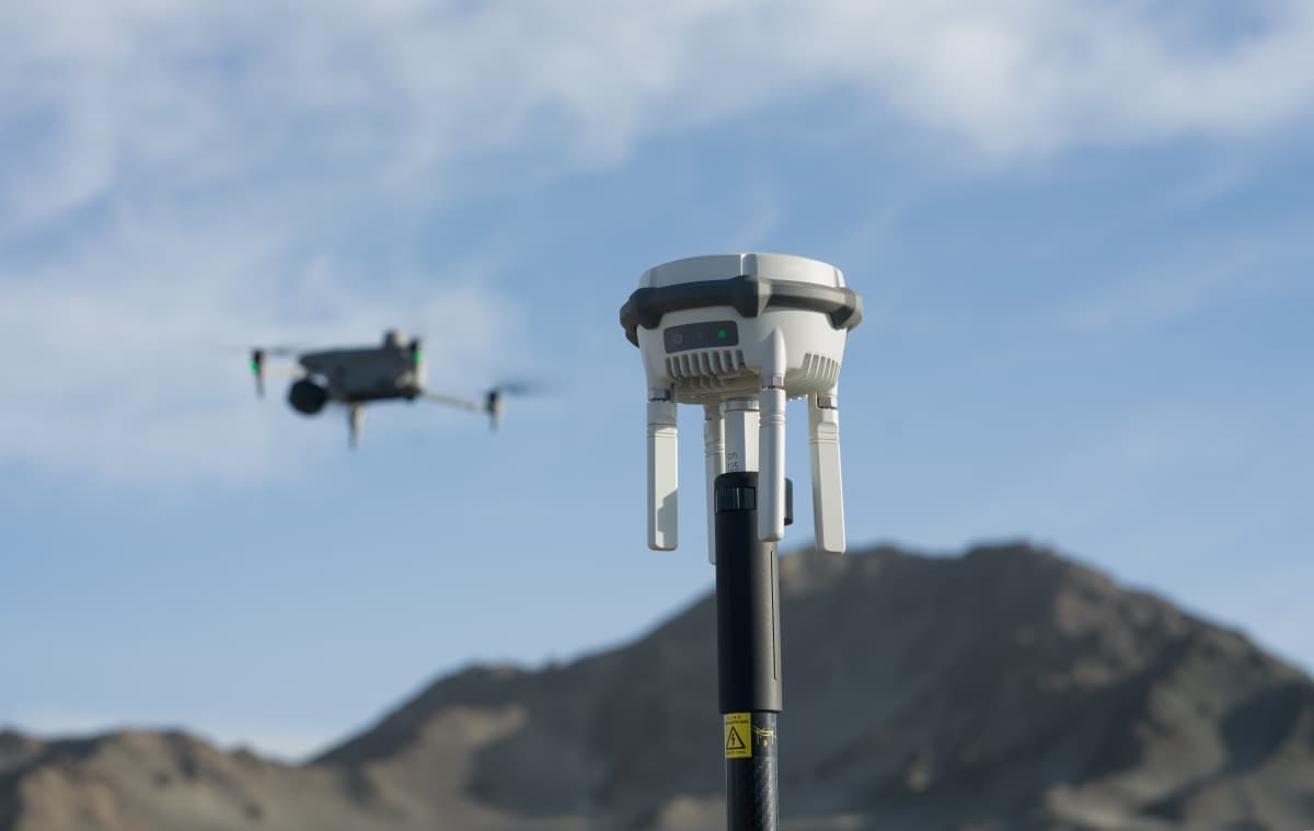 "En drönare flyger nära en GPS-mottagare på en stång, med berg i bakgrunden under en klar himmel."