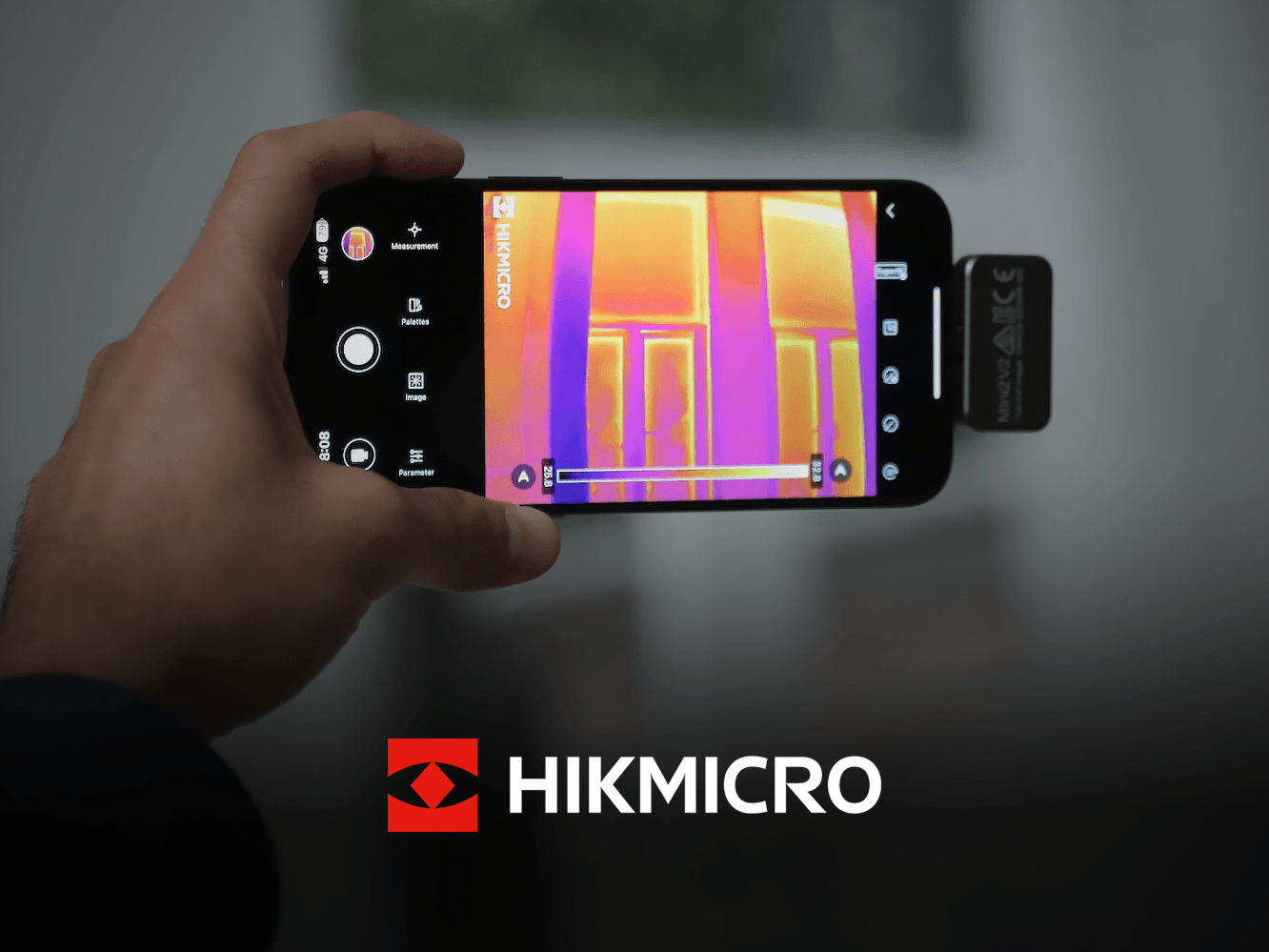 En hand håller en smartphone med en värmekamera som visar en termisk bild av ett rum, med HIKMICRO-logotypen synlig.