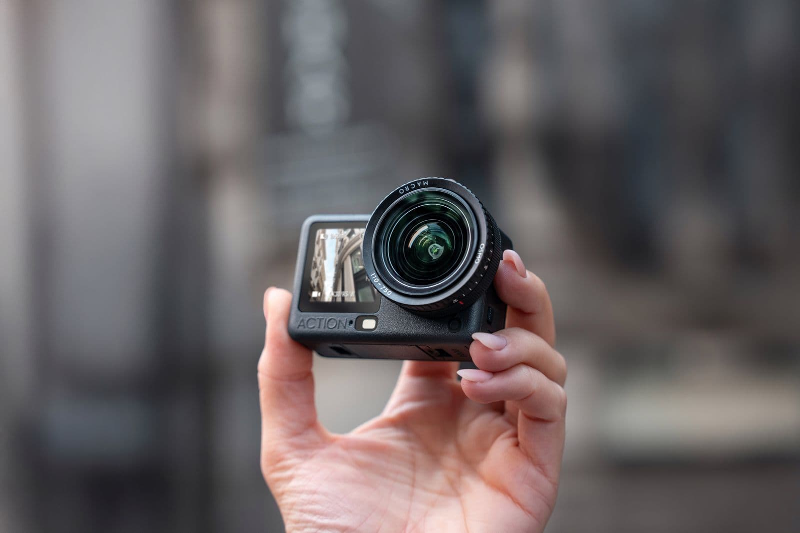 En hand håller en kompakt actionkamera med ett stort objektiv, mot en suddig bakgrund.