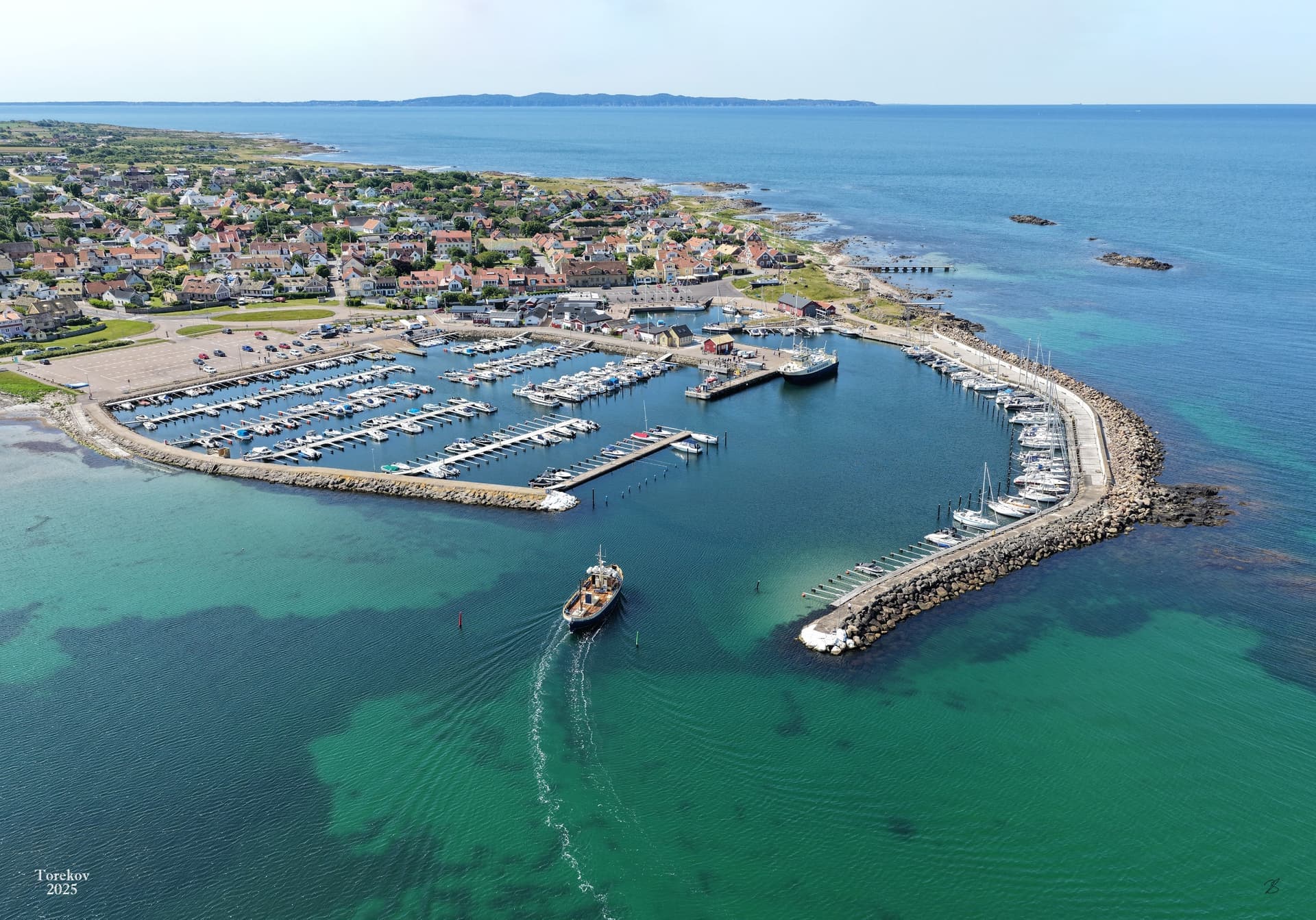 En pittoresk hamn med båtar förtöjda vid bryggor, omgiven av en kuststad och det öppna havet i bakgrunden.