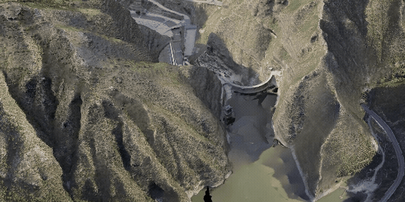 En drönarvy visar en damm inbäddad mellan branta bergssluttningar, med vatten som samlas nedanför.