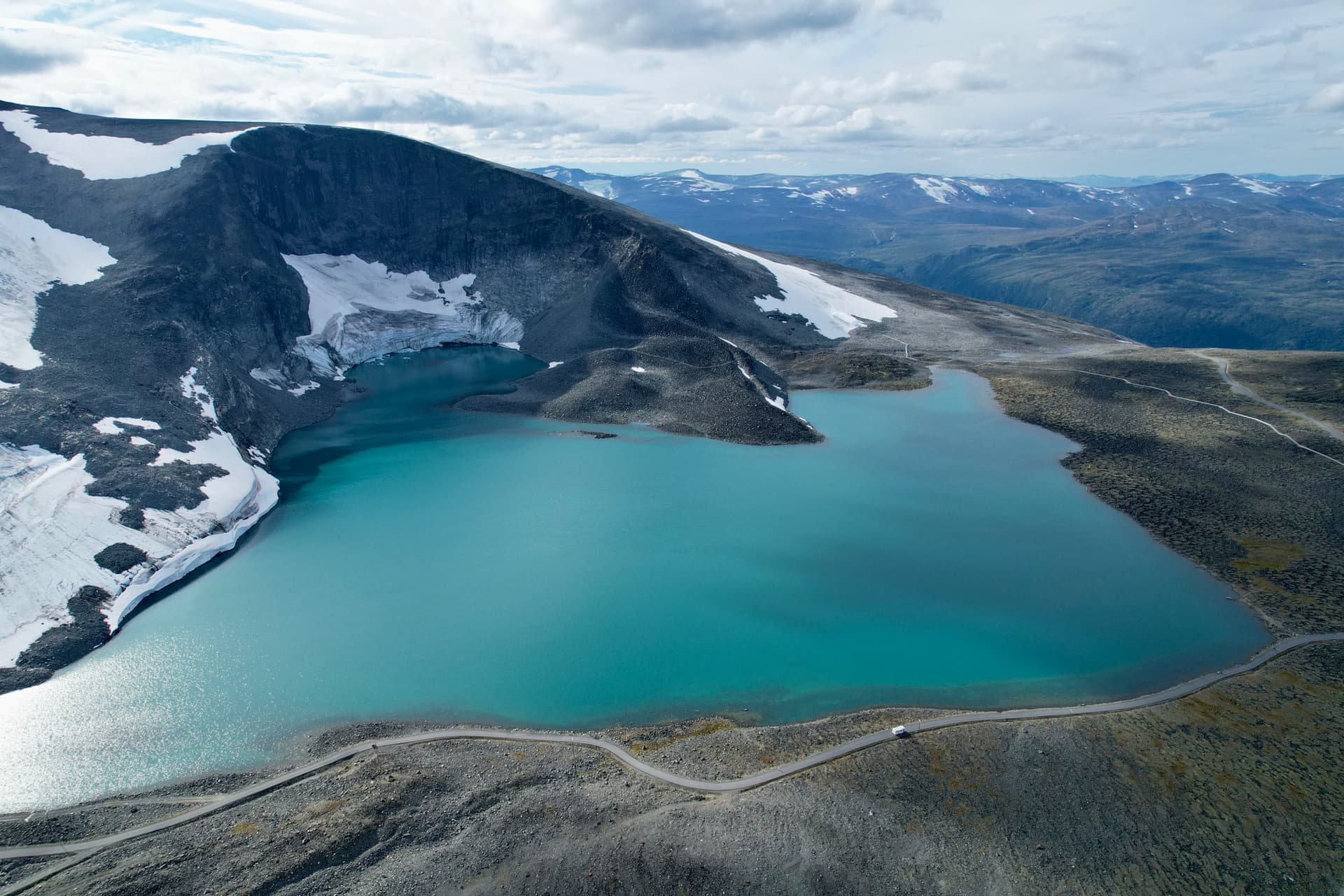 En turkos sjö omgiven av snötäckta berg och en slingrande väg i ett dramatiskt fjällandskap.