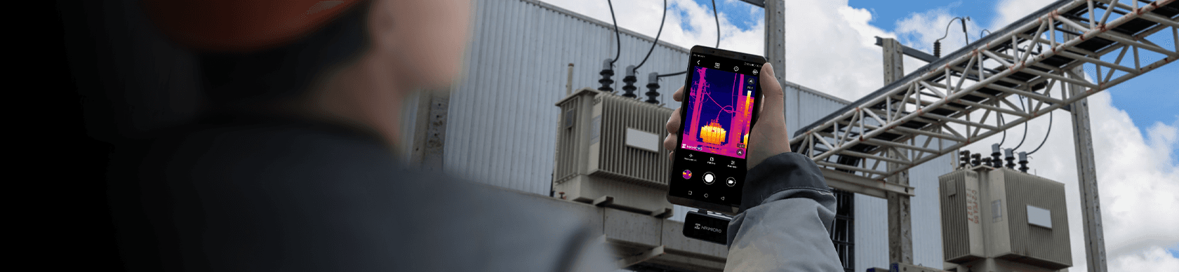 En person använder en smartphone med värmekamera för att inspektera elektrisk utrustning utomhus.
