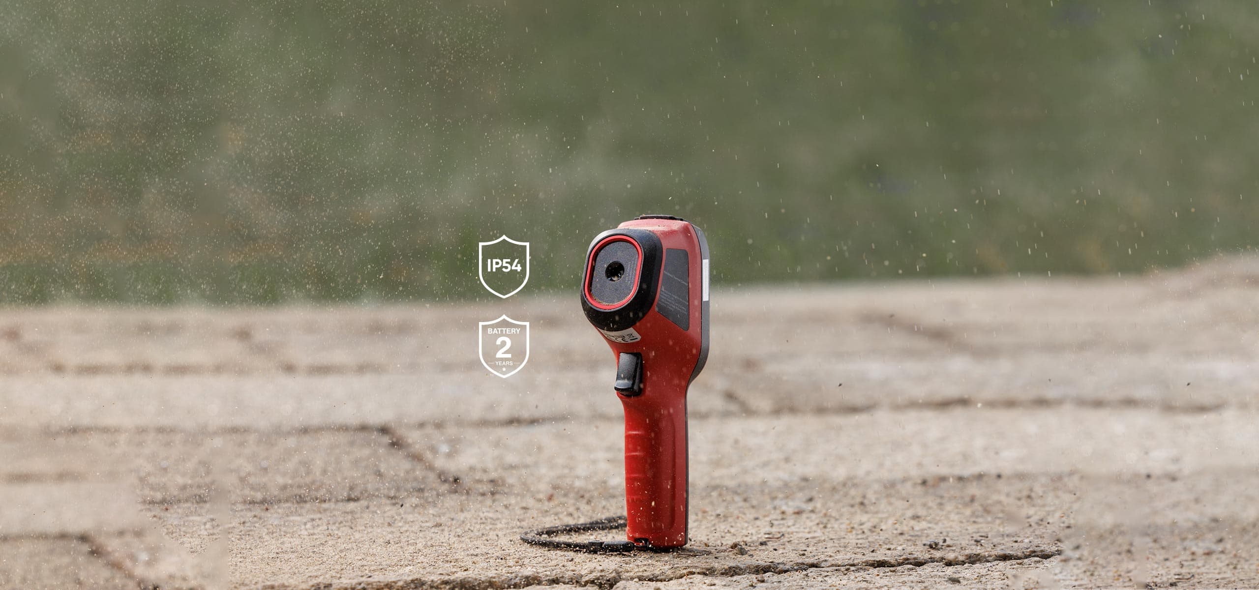 En robust, röd enhet står på en våt yta, omgiven av regndroppar, med IP54- och tvåårig batterisymboler i bakgrunden.