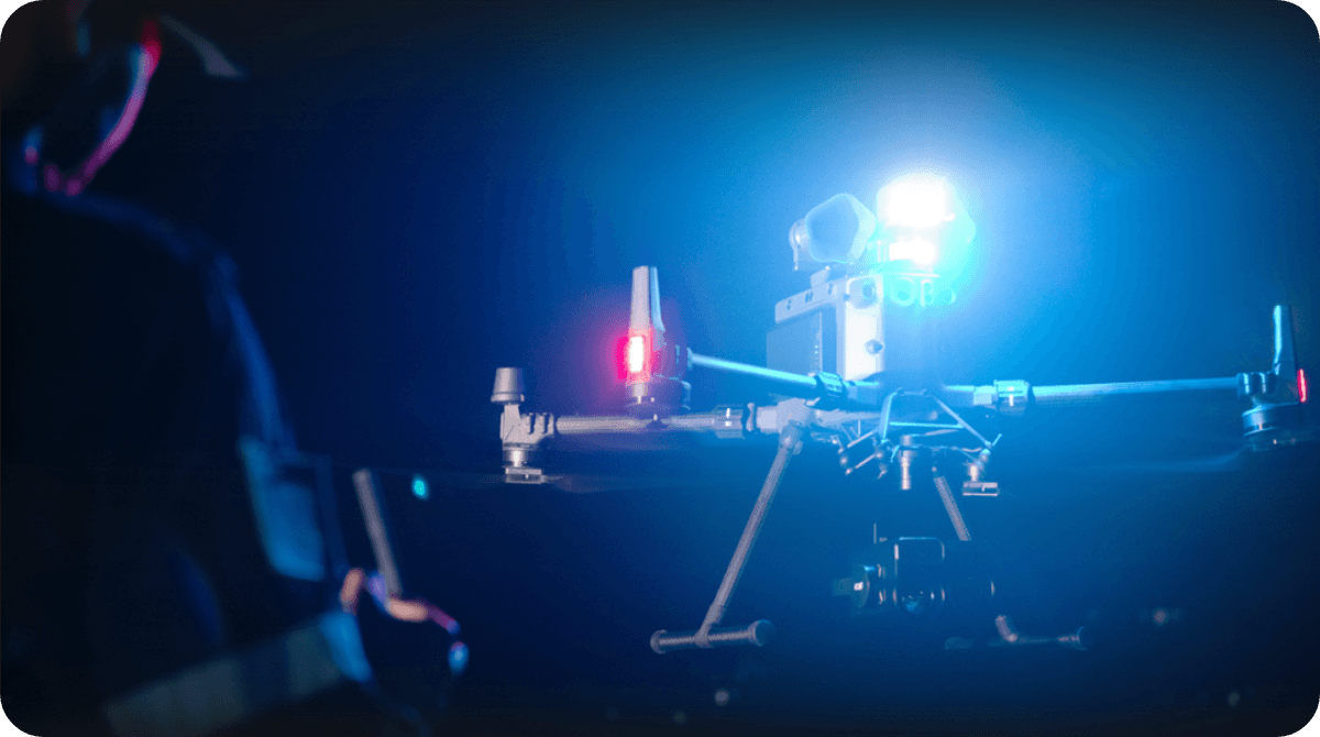 En drönare med lysande blå och röda lampor svävar i mörkret, styrd av en person i bakgrunden.