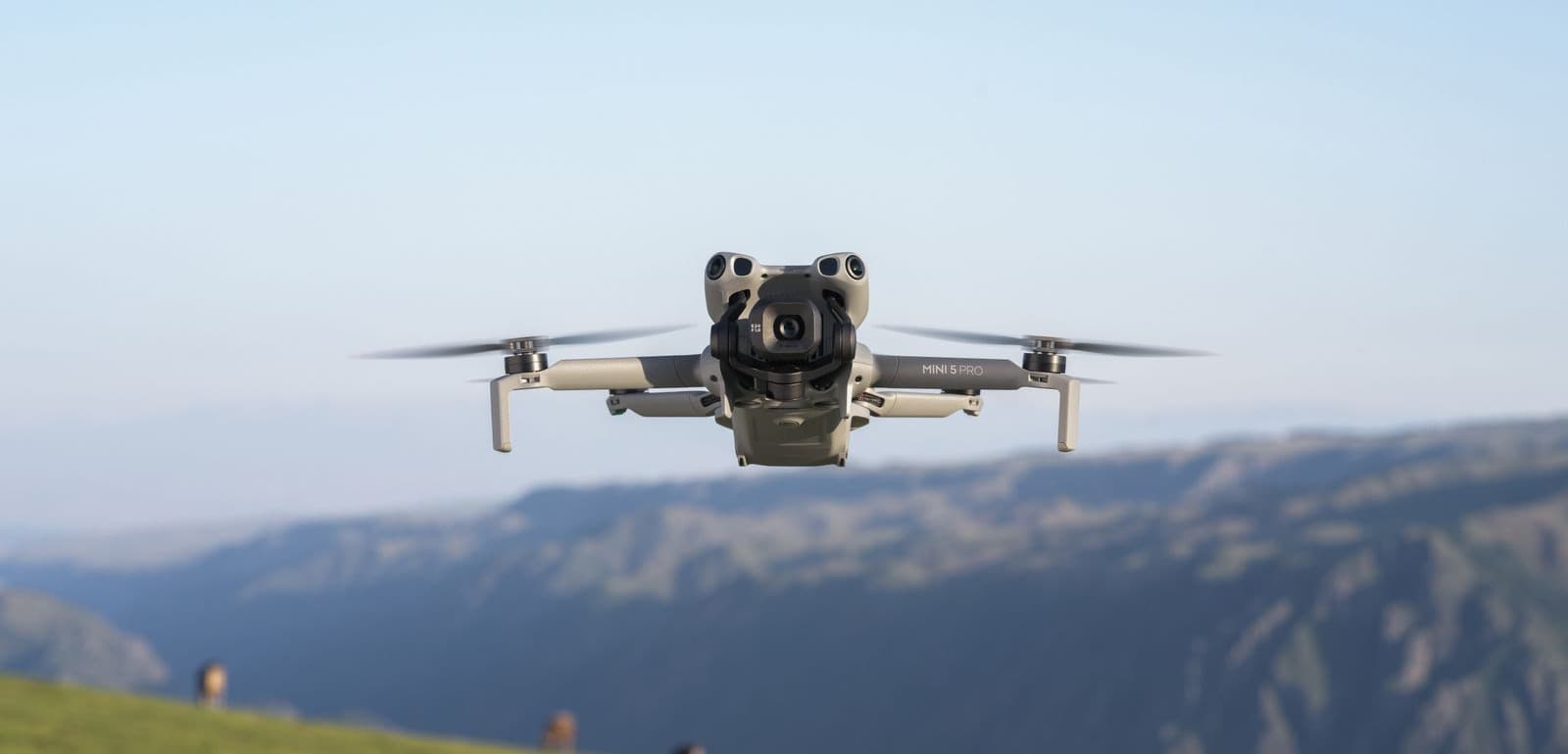 En drönare svävar över ett naturskönt landskap med berg i bakgrunden under en klarblå himmel.