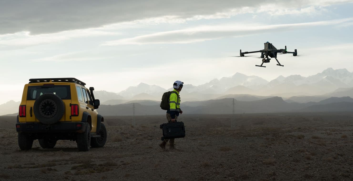 En person i skyddsutrustning och varselställ står bredvid en gul terrängbil och övervakar en DJI Matrice 400-drönare som flyger över ett landskap med berg i bakgrunden.