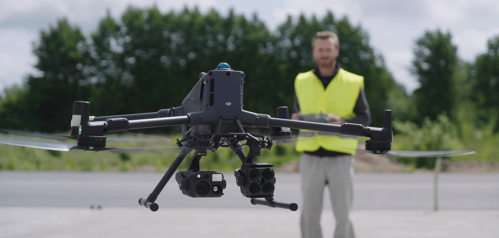 En Matrice 400 / M400 svävar i luften medan en person i gul varselväst styr den med en fjärrkontroll i en utomhusmiljö.