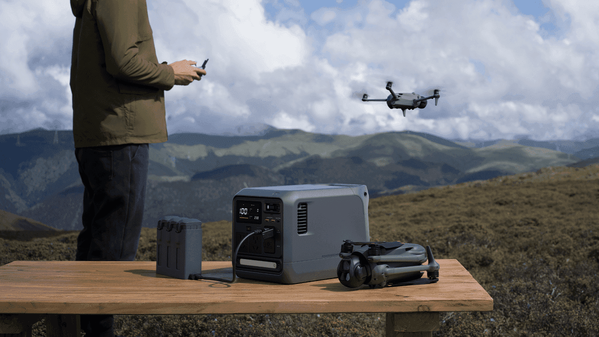 En person styr en drönare i en naturskön bergsmiljö, med en bärbar kraftstation (DJI Power 1000 Mini) och drönartillbehör på ett träbord.
