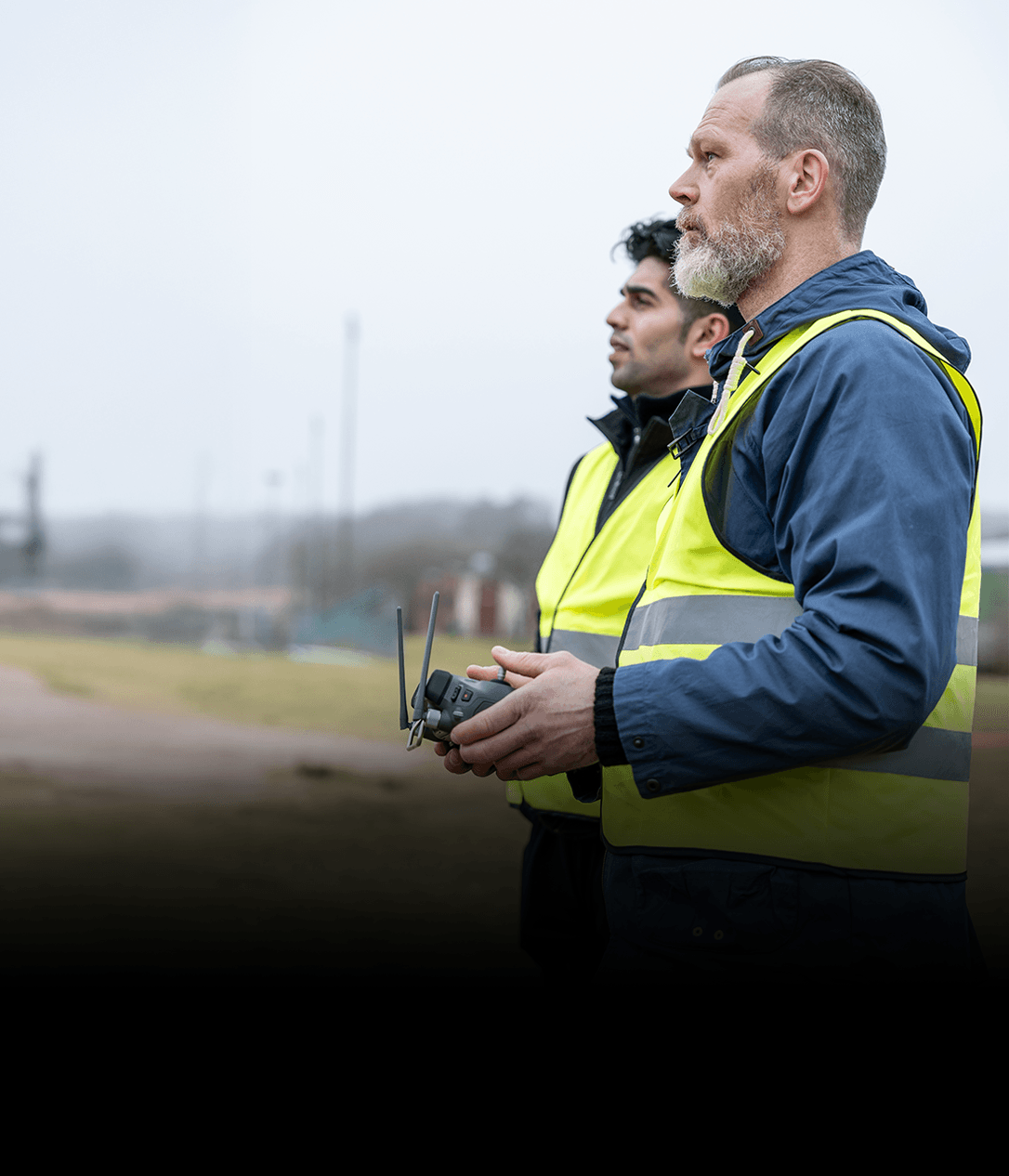 Två personer i reflexvästar står utomhus och styr en drönare med en fjärrkontroll.