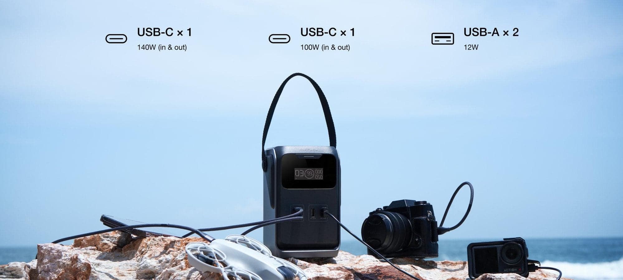 En bärbar kraftstation med flera USB-portar laddar en drönare, kamera och actionkamera på en stenig strand med havet i bakgrunden.