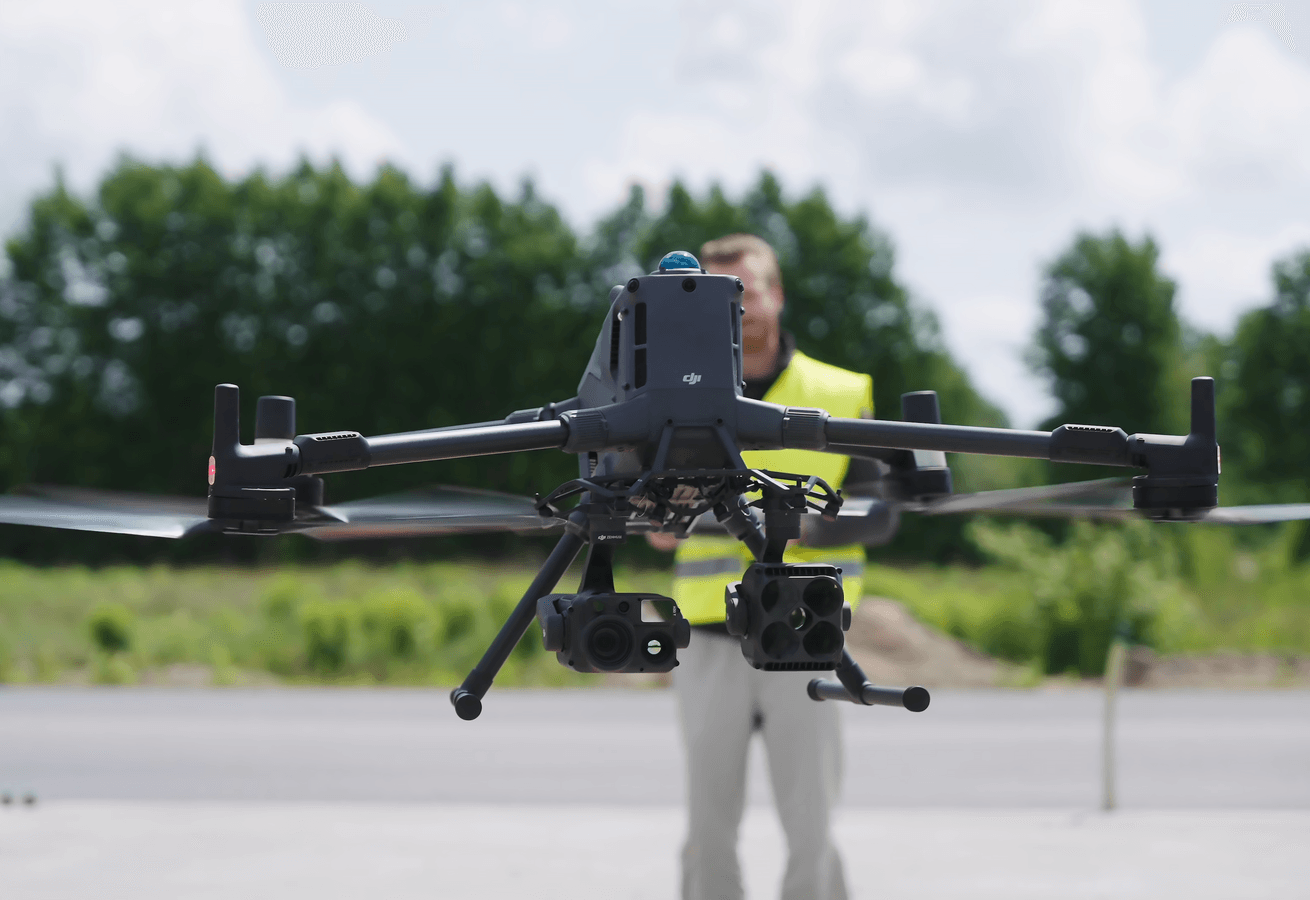 Drönare för inspektion: En DJI Matrice 400-drönare med en monterad DJI Zenmuse H30T-kamera svävar framför en person i en gul varselväst på en öppen plats med träd i bakgrunden.