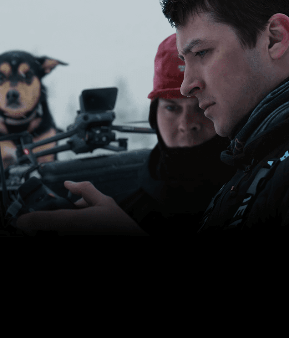 En person fokuserar på att styra en drönare i en snöig miljö, medan en hund observerar i bakgrunden.
