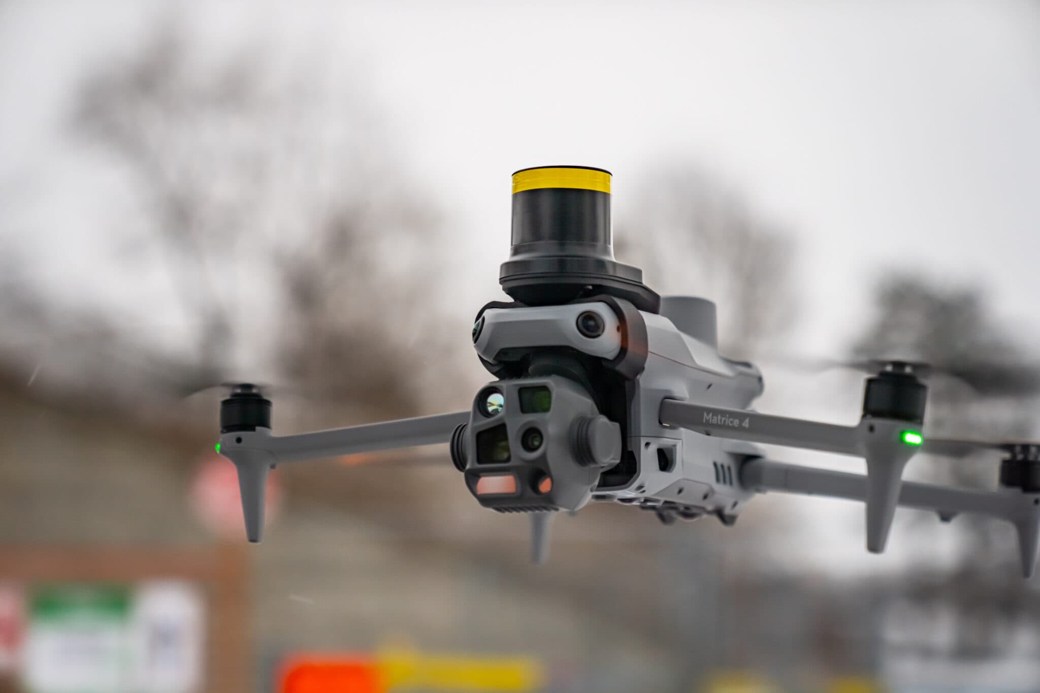 En drönare svävar i luften med snurrande propellrar, mot en suddig bakgrund av träd och byggnader.