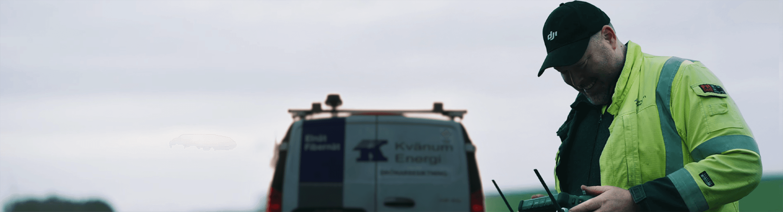 En person i en gul skyddsjacka använder en fjärrkontroll bredvid en servicebil på en öppen plats.