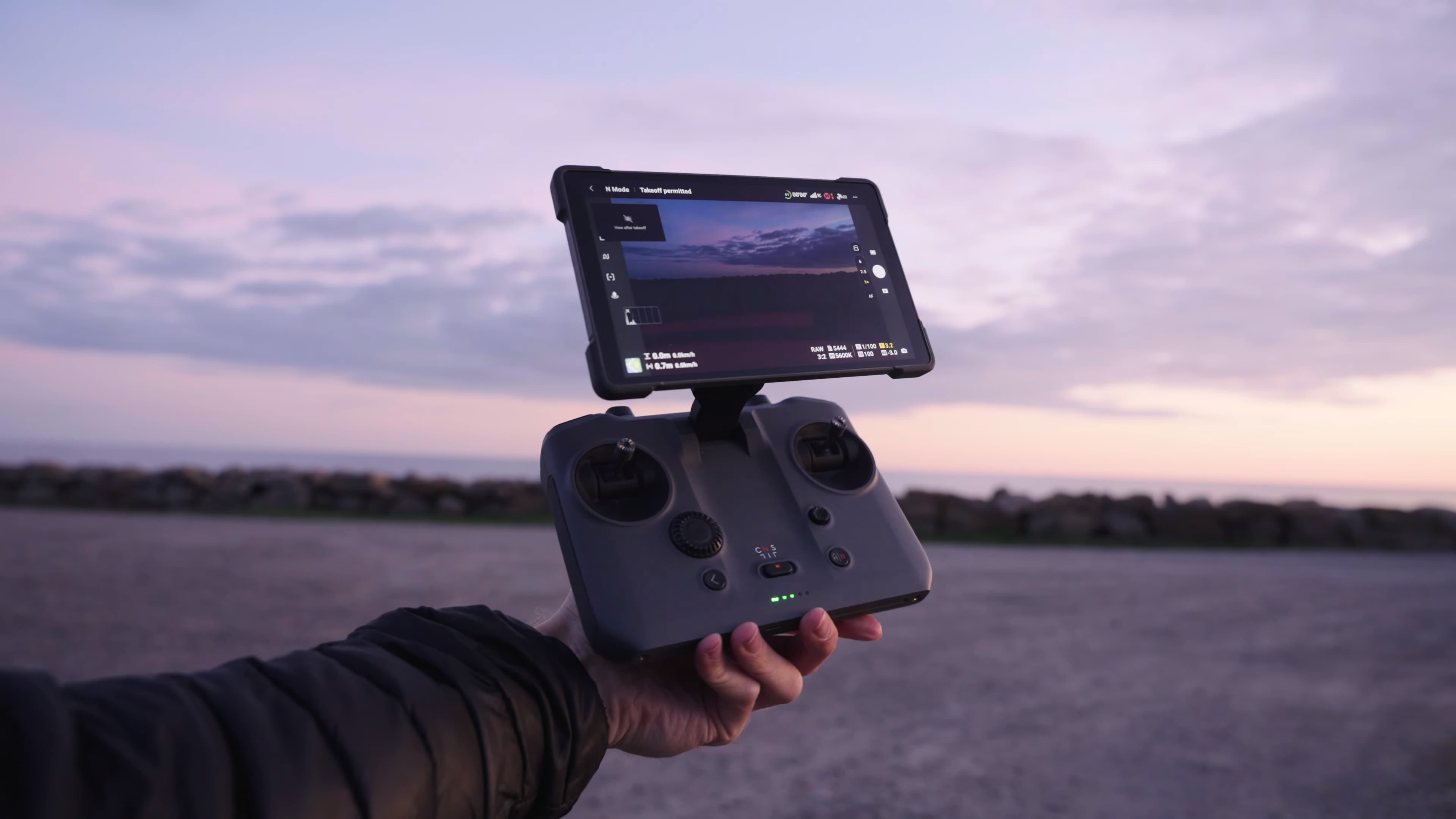 En person håller en drönarkontroll med en skärm som visar en vy av landskapet vid solnedgången.