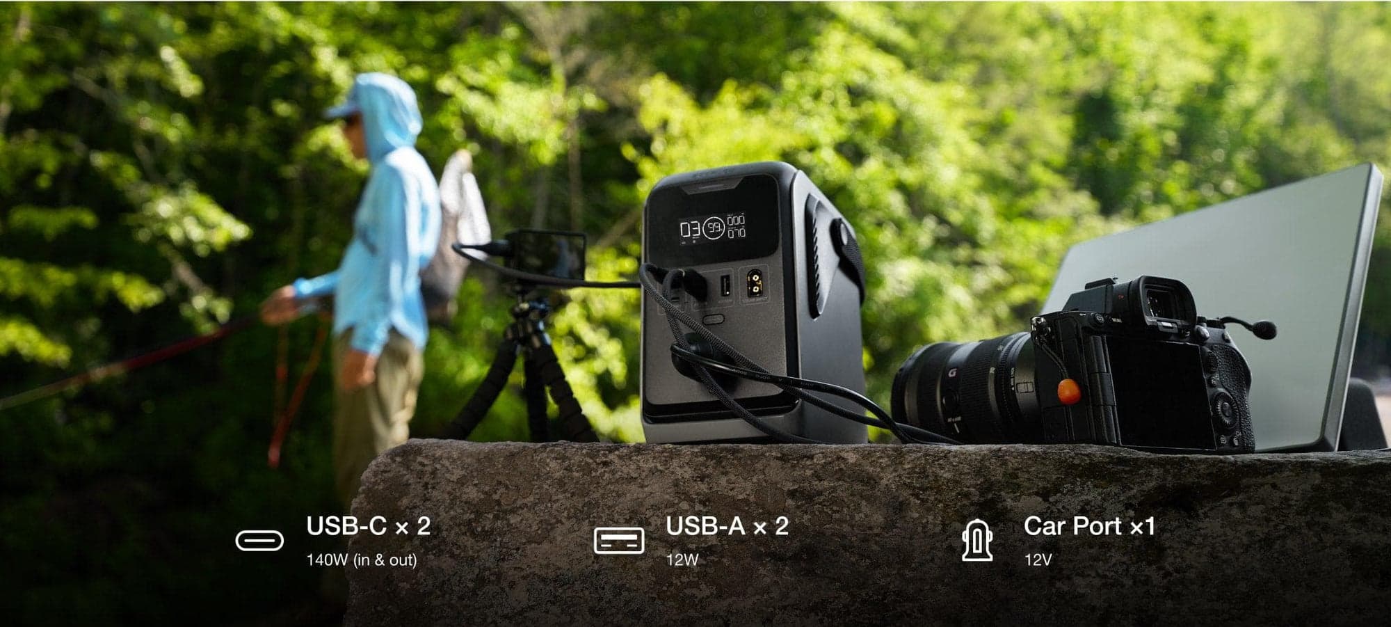 En bärbar kraftstation med flera portar står på en sten, omgiven av en kamera och en laptop, medan en person fiskar i bakgrunden.