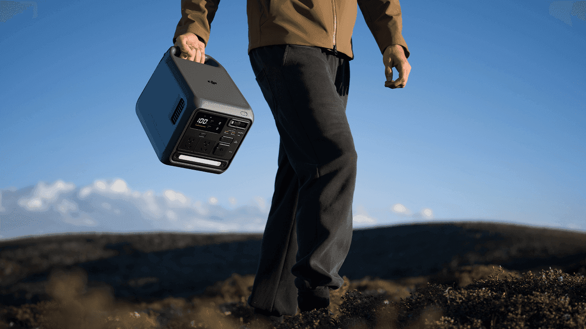 En person bär en portabel kraftstation (DJI Power 1000 Mini) över ett öppet landskap under en klarblå himmel.