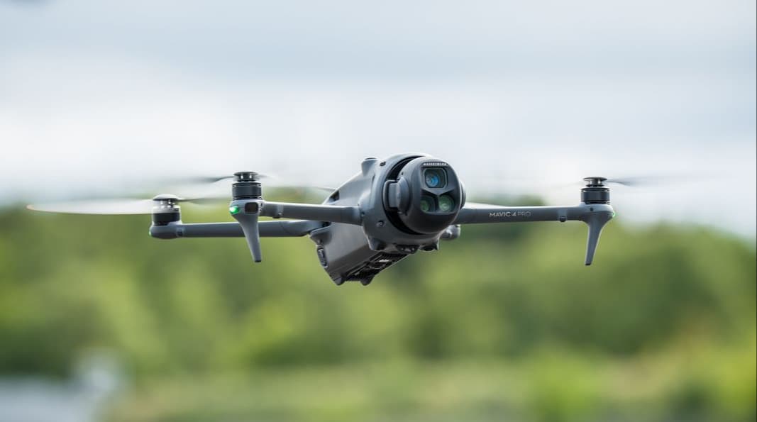 En Mavic 4 Pro svävar i luften mot en suddig bakgrund av grönska och himmel.