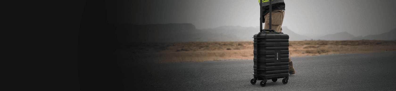 En person står på en öde väg med en robust svart resväska på hjul, omgiven av ett kargt landskap.