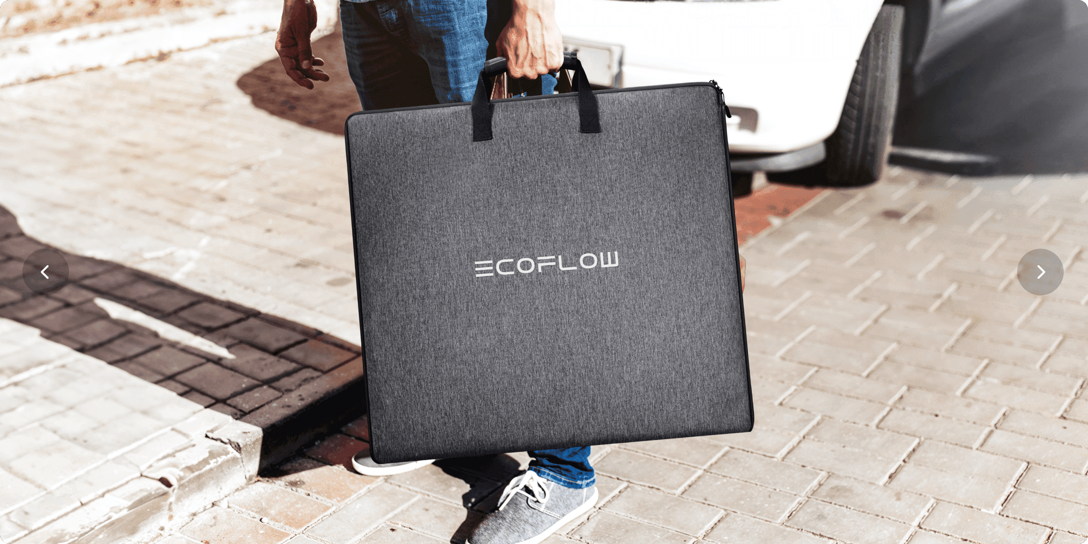 En person bär en stor, grå EcoFlow-väska på en trottoar bredvid en parkerad bil.