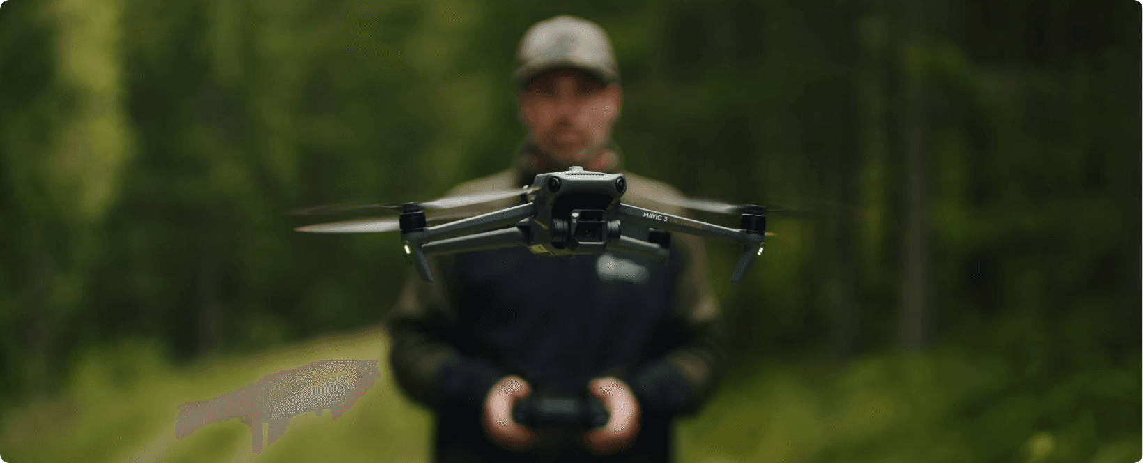 En drönare svävar i luften framför en person som håller en fjärrkontroll, omgiven av en skogsmiljö.