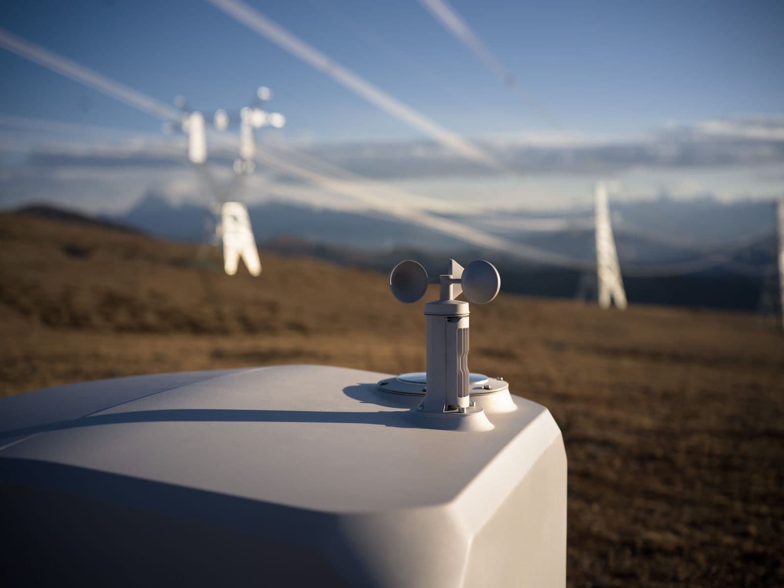 En väderstation med en vindmätare står på en kulle, med kraftledningar som sträcker sig över landskapet i bakgrunden.