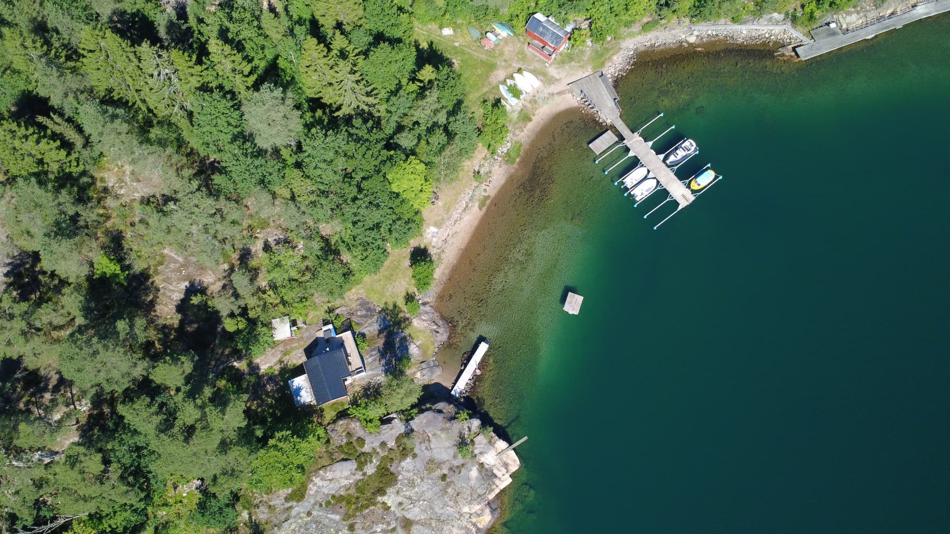 Flygbild av en brygga med båtar vid en skogskantad kustlinje, med en stuga och grönska i närheten.