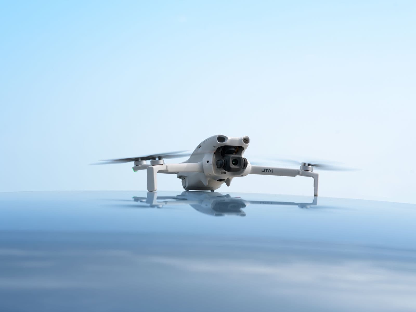 En vit drönare med snurrande propellrar svävar över en reflekterande yta mot en klarblå himmel.