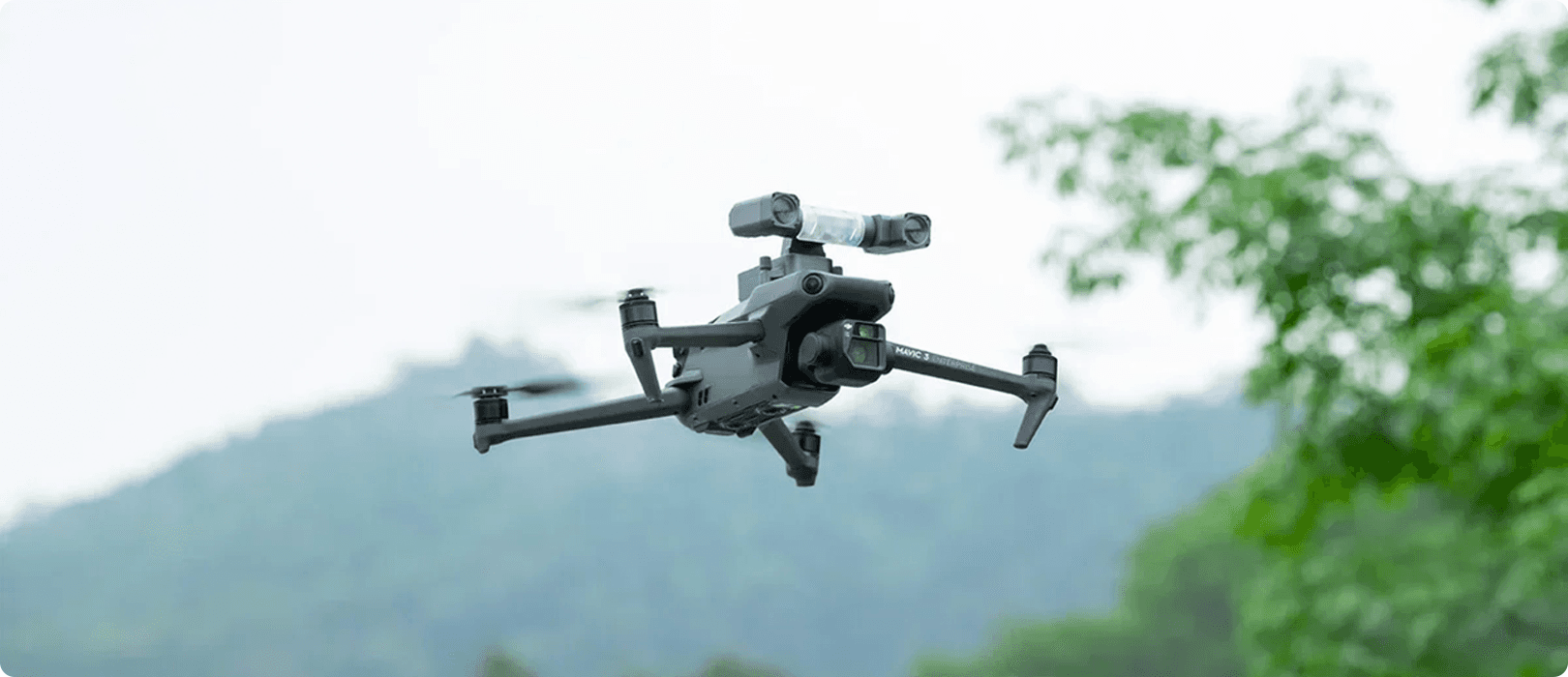 En DJI Mavic 3E-drönare utrustad med drönartillbehör från CZI svävar i luften mot en bakgrund av träd och dimmiga berg.