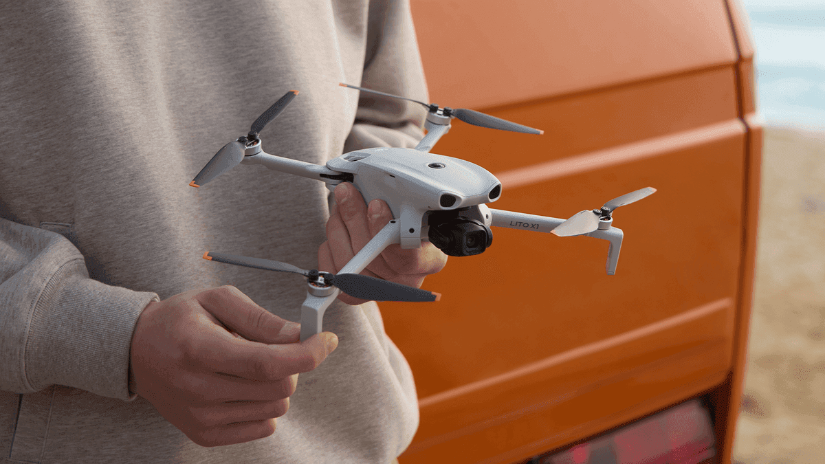 En person håller en kompakt DJI Lito X1-drönare framför en orange bil, redo för flygning vid stranden.